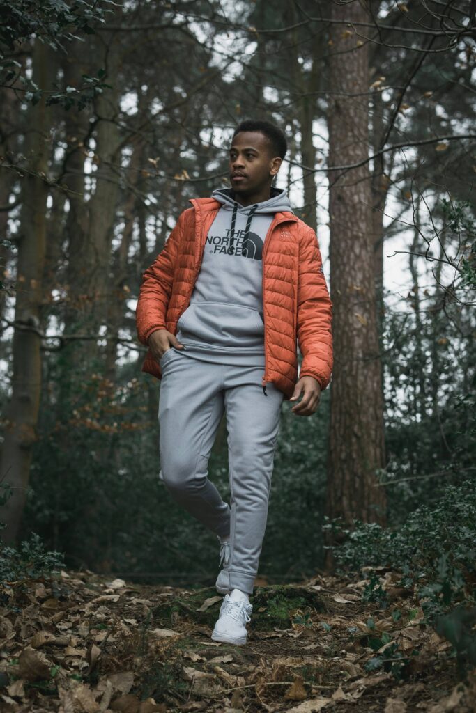 Un homme marchant dans une forêt, portant une tenue de sport grise avec une veste orange et des baskets blanches, entouré d'un environnement naturel calme avec des arbres et des feuilles tombées.