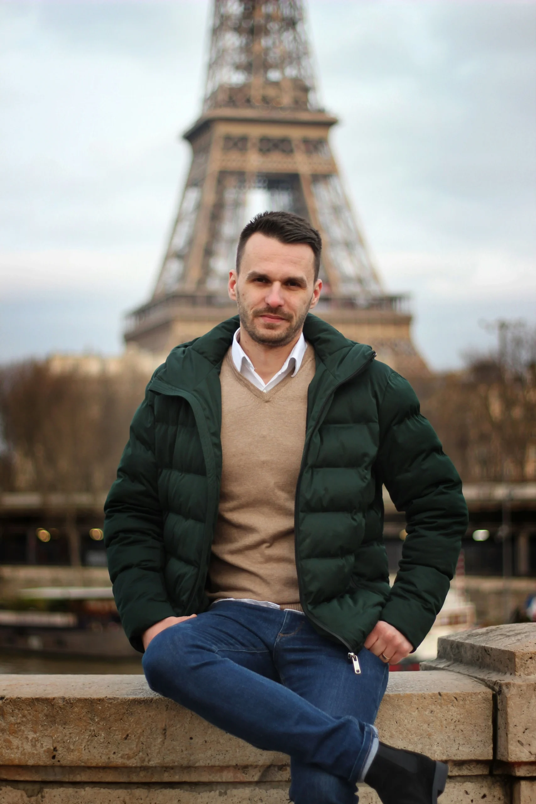 Un homme assis devant la tour Eiffel, portant une veste verte, un jean bleu et une chemise blanche avec un pull beige, dans une ambiance calme à Paris.