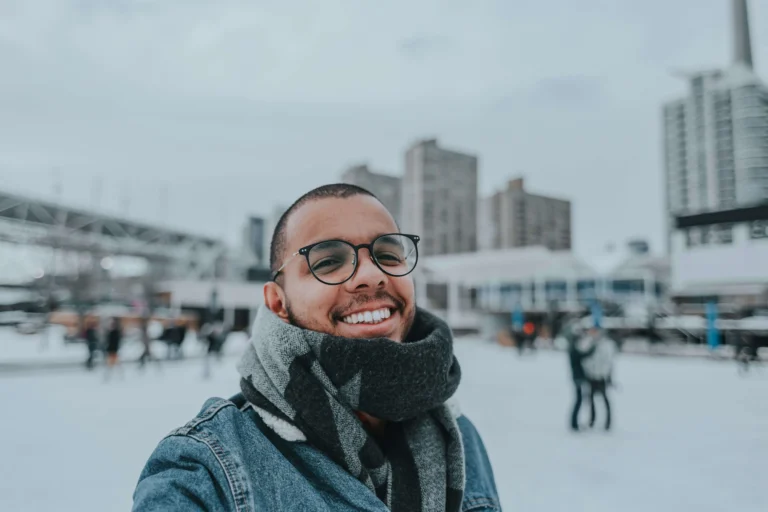 Un homme souriant portant des lunettes, une écharpe grise et une veste en jean, prenant un selfie en extérieur dans un cadre urbain enneigé.