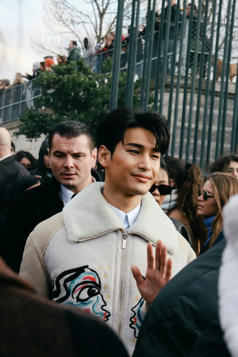 Homme souriant portant une veste beige avec des motifs, saluant dans une foule.