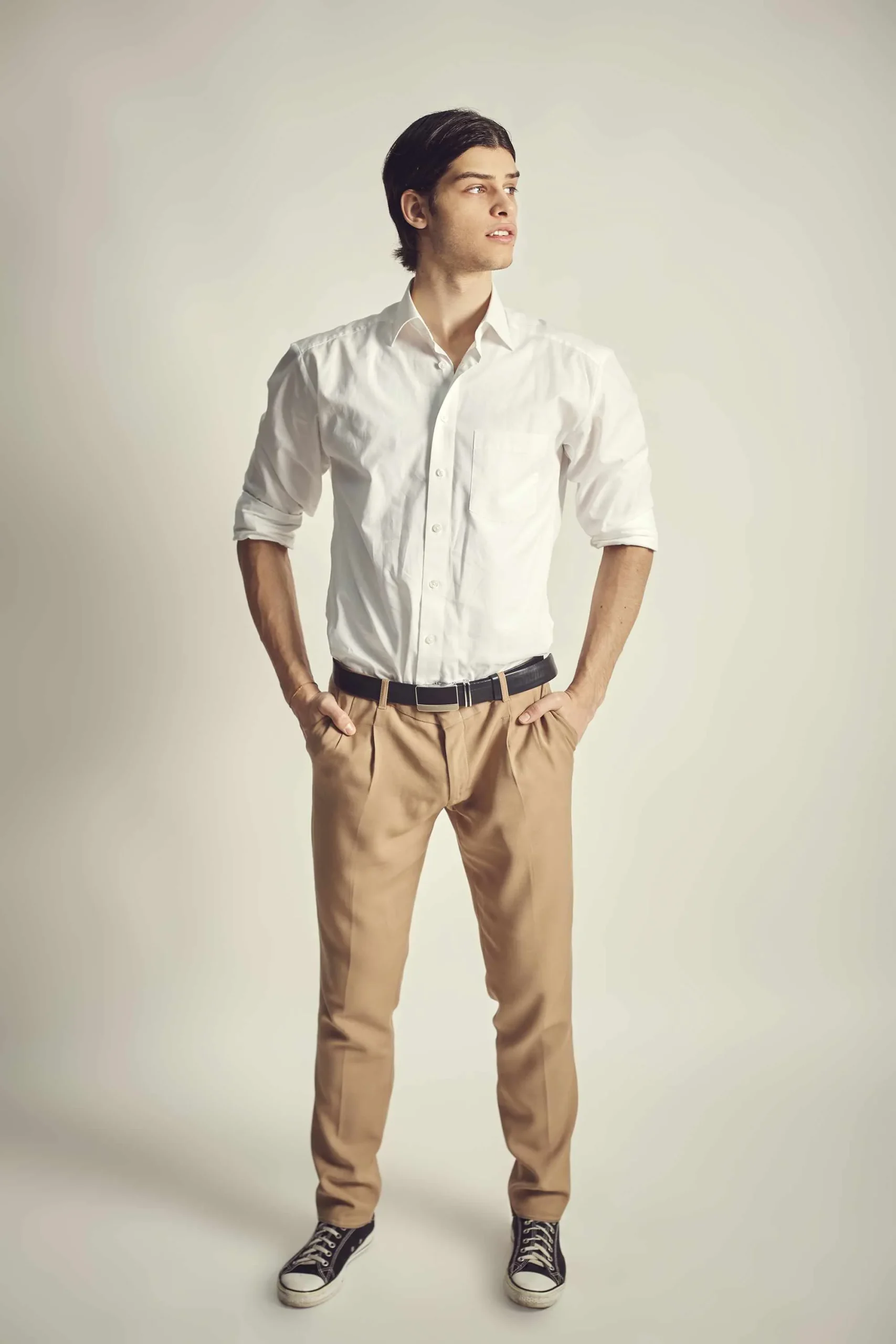 Homme debout en chemise blanche et pantalon beige, mains dans les poches, portant des baskets noires.